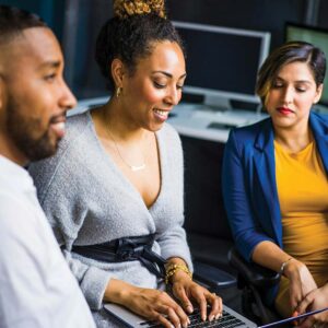 Three employees sit together as one types into her laptop as they discuss a project.